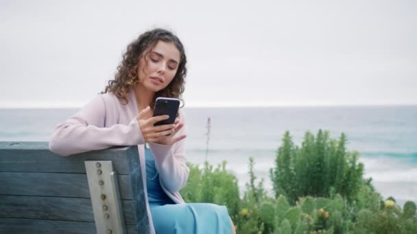 微笑的年轻女性使用智能手机装置 红色相机 在社交媒体上回答信息 快乐卷曲的女孩坐在海洋公园的木制长椅上 在手机屏幕上在线聊天 — 图库视频影像