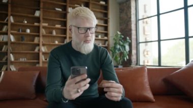 Smiling smart man in eyeglasses purchasing online 4K. Happy male shopper using instant easy mobile payments. E-banking app service, customer holding credit card and smartphone sitting on couch at loft