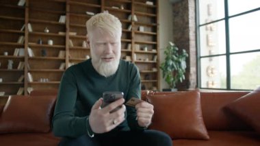 Smiling young albino man holding credit card and smartphone sitting on couch at home. Happy male shopper using instant easy mobile payments. E-banking app service for customer purchases online store