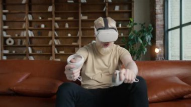 VR gözlüklü sanal oyuncular modern çatı katı tarzı dairelerde sanal gerçeklik oyunu oynarlar. Hippi sakallı adam oyun konsepti. Genç yakışıklı adam etrafına bakıyor ve joystick kullanarak sanal silah ateşliyor.