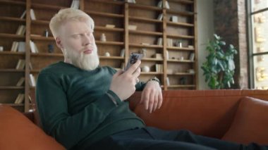 Close up view of serious middle aged man with blond hair sitting on leather sofa using phone in modern urban loft apartment, texting message scrolling news. Technology lifestyle slow motion dolly shot
