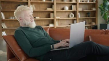 Modern daire 4K 'de uzaktan daktilo yazan konsantre sarışın albino girişimci. Ev ofisinin oturma odasındaki kanepede dizüstü bilgisayar kullanarak milenyum adamı olmaya odaklanmış.