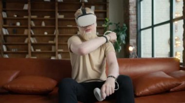 Sanal gerçeklik kulaklığı kullanan hippi bir gencin sinematik portresi. Sarışın sakallı adam evdeki koltukta oturuyor ve ağır çekimde video oyunları oynayarak el hareketi yapıyor.
