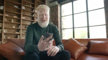 Discolored white haired man with albinisms disease causing neurological disorder with involuntary head shaking trembling. Portrait of Albino guy taking by video call using front camera on smartphone