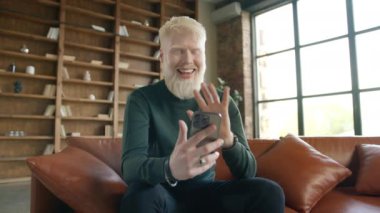 Excited active guy in casual elegant outfit using mobile phone. Technology lifestyle use concept. Smiling young blond man talking video conference call using smartphone in living room at modern home