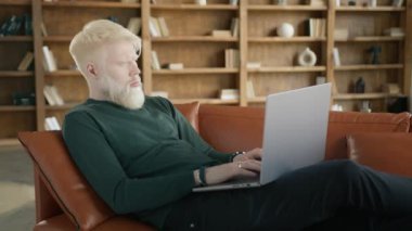 Ev ofisinin oturma odasındaki kanepede dizüstü bilgisayar kullanarak milenyum adamı olmaya odaklanmış. Yoğunlaşmış sarışın albino girişimci modern daire 4K yavaş bir şekilde defter üzerinde çalışıyor.