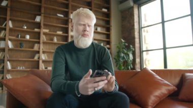 Focused man with blond hair frowning brows while sitting on leather sofa using phone. Serious concentrated Albino guy scrolling news, reading emails in modern urban loft apartment on motion background