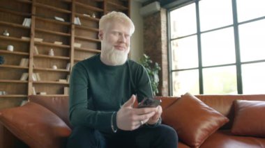 Positive guy using apps playing mobile game chatting in social media surfing web on phone in modern home. Young smiling albino man holding smartphone device texting sms message sitting at home office