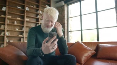 Smiling young blond man talking video conference call using smartphone in living room at modern home. Excited active guy in casual elegant outfit using mobile phone. Technology lifestyle use concept
