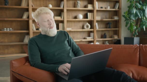 ポジティブなミレニアル世代のフリーランサーは 自宅のオフィスのリビングルームでソファーにノートパソコンを使用しています ブロンドアルビノの起業家はひげに触れ 近代的なロフトアパート4Kの遅いノートブックにタイピング — ストック動画