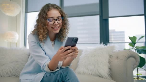 用手机用时髦眼镜微笑迷人的女性 中关闭慢动作红色相机拍摄 漂亮的女人坐在沙发上 在现代设计公寓的内部使用智能手机 — 图库视频影像
