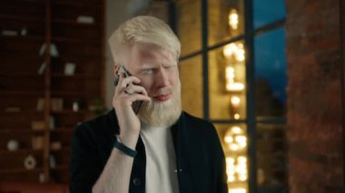 Gülümseyen albino girişimci gece yarısı çatı katındaki ofiste akıllı telefondan konuşuyor. İş arkadaşıyla telefon görüşmesi yapan sıradan bir iş adamının portresi. Şaşırmış yüz ifadesi olan hippi adam. 