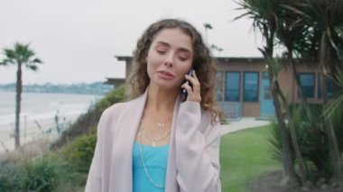 Bulutlu bir yaz gününde okyanusta cep telefonuyla konuşan mutlu bir kadın. Gülen kıvırcık kadın şık bir hırka ve mavi ipek elbise giyiyor yeşil parkın yanından geçiyor ve akıllı telefon kullanıyor.