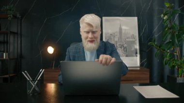 Mutlu albino işadamı ofisteki dizüstü bilgisayarla işini bitiriyor. Erkek girişimci iş gününün sonunda eğleniyor. Pozitif yönetici masadaki bilgisayarı kapatıyor. Ofis sandalyesinde rahatlayan gülümseyen adam.