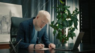 Odaklanmış albino işadamı portresi iş yerinde doküman dolduruyor. Erkek girişimci ofiste dizüstü bilgisayarla çalışıyor. Kendine güveni tam bir çalışan, kağıtlarda kalemle notlar alıyor. 