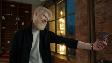 Bencil albino adam, gece sokak tabelasında çatı katındaki stüdyoda akıllı telefondan selfie çekiyor. Adam selfie çekmek için cep telefonu kullanıyor. Sosyal medya blogu için Freelancer hipster blog yazarı, yaşam tarzı