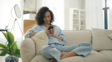 Gerçek insanların yaşam tarzı akıllı telefon eğlencesi 4K. Gülümseyen Afro-Amerikan tutkulu kadın elinde akıllı telefon tutuyor. Çift ırklı kız cep telefonu kullanıyor. Sosyal medya izliyor. Gün ışığında evde oturuyor.