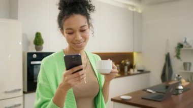 Çeşitli model kıvırcık kadın kahve içiyor ve elinde akıllı telefon tutuyor. İnternetten yemek siparişi veriyor, ürünleri yavaş çekimde seçiyor. Modern mutfakta akıllı telefon kullanan genç ve güzel bir model.