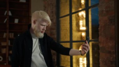 Kaliteli albino adam stüdyoda akıllı telefondan selfie çekiyor. Şehir gece hayatı konsepti. Kapalı alanda cep telefonu kamerası kullanan gülümseyen bir hippi. Modern cihaz 4K 'da özçekim yapan yakışıklı bir adam.