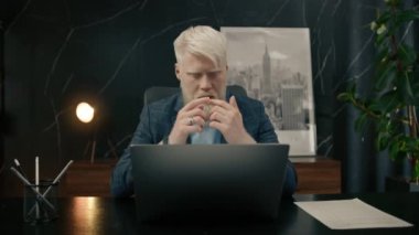 Düşünceli, genç, beyaz, harmanlanmış albino iş adamı ofiste dizüstü bilgisayarla oturup ilham almayı, maliyeti hesaplamayı, arama problemi çözümü fikirlerini, düşünce konseptinde kaybolmayı hayal ediyor.