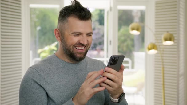 Podekscytowany Uśmiechnięty Człowiek Stojący Domu Czytający Maile Przez Telefon Dobrymi — Wideo stockowe