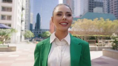Modern şehir manzarasının ortasında yeşil ceket giyerek hareketli arka planda dururken büyüleyici bir şekilde gülümseyen alımlı iş kadınının portresi. Mutlu kadın hayattan zevk alıyor.