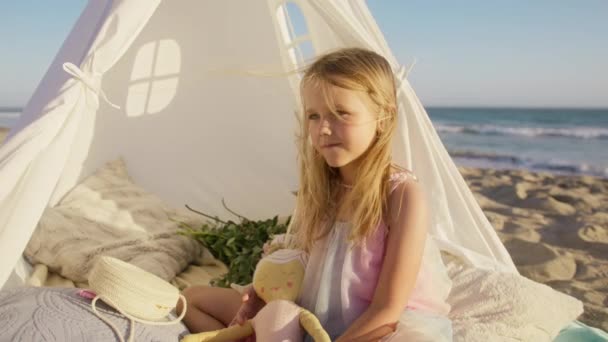 小さな幸せな少女は 手の詰まった人形のおもちゃを保持する興味深い物語を話します ゴールデンサンセットライトの海のビーチで愛らしい子供の面白い顔 風4Kで飛んでいるロングブロンドの髪の可愛い少女 — ストック動画