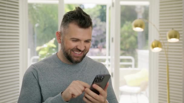 モバイル勝利コンセプト はい手によるジェスチャー 良いニュースを読むSmsメッセージを受け取る驚くべき幸せな男 自宅で現代的なデザインでスマートフォンを見る成功を祝う興奮した男性の勝者 — ストック動画