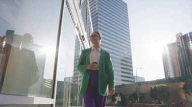 Gülümseyen çekici iş kızı gün ışığının tadını çıkarıyor. Kariyer sahibi insanlar 4K 'yi vurdu. Başarılı genç kadın kahve fincanı günbatımında kentsel arka planda modern iş binalarında yürüyor.