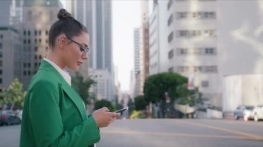Resmi yeşil takım elbise ve beyaz tişört giyen gülümseyen Amerikalı kadın şehir merkezindeki iş yerlerinin önünde cep telefonuyla mesaj yazıyor. Cep telefonu akıllı telefon ağır çekim 4K USA