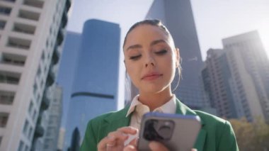 Mutlu bir şekilde gülümseyen, takma kirpikli, akıllı telefondan yazı yazan çekici bir kadın. Düşük açılı, sinematik bir bebek resmi. Modayı takip eden şık bayan işten sonra güneşli bir günde arka planda mercek ışığıyla eğleniyor.