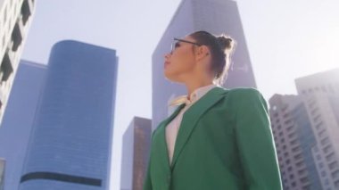 Güzel modern yönetici ağır çekimde. Kendine güvenen, resmi kıyafetli genç bir kadın modern şehir merkezinin finans bölgesinde sokaklara bakıyor. İş kadını portresi düşük açılı.