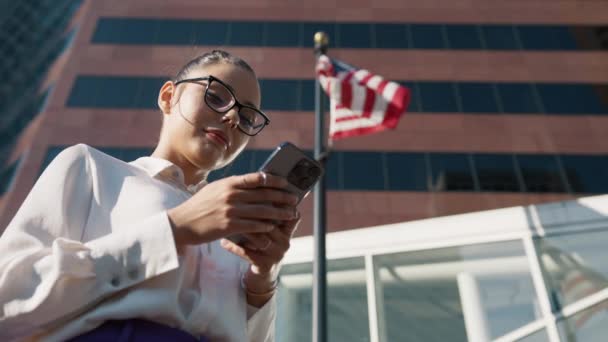 近代的なオフィスビルの近くでスマートフォンを閲覧するビジネスマンは フラグポールに風を吹くアメリカの旗を掲げています 携帯電話でアプリを使用して正式な着用で魅力的な女性 アメリカンコンセプト4K — ストック動画