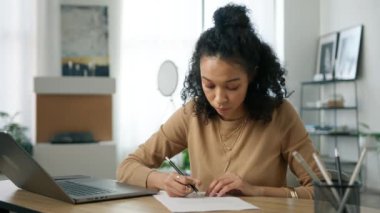 Yoğunlaştırılmış odaklanmış Afrikalı kız öğrenci iş belgelerine not alıyor, dizüstü bilgisayarına bakıyor. Yurt içi ofis masasında uzaktan çalışan siyahi genç bir kadın ve İnternet sitesi 4K 'den öğreniyor.