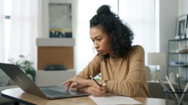 Merkez büroda uzaktan çalışan çok ırklı genç bir kadın dizüstü bilgisayara bakıyor, İnternet sitesinden öğreniyor. Yoğunlaştırılmış odaklanmış Afrikalı kız öğrenci iş belgesi 4K 'ye notlar alıyor.