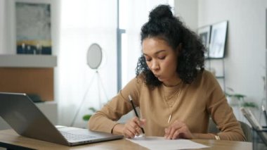 Yurt içi ofis masasında uzaktan çalışan ve internet sitesi 4K 'de öğrenen siyahi genç bir kadın. Yoğunlaştırılmış odaklanmış Afrikalı kız öğrenci iş belgesine notlar alıyor, dizüstü bilgisayarına bakıyor.