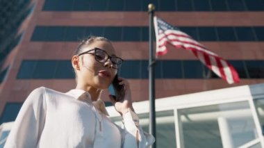 Gülümseyen kadın, şehir merkezindeki iş yerinde rüzgarda dalkavukluk eden büyük Amerikan bayrağı altında telefon görüşmesi yapıyor. Politika, Ekonomi, İş Konsepti. Düşük açılı sinematik ağır çekim.