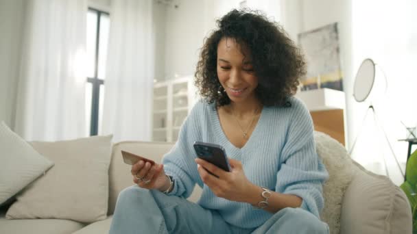 スマートフォンアプリ4Kで購入する笑顔のお客さん クレジットカードを保持するカーリーヘアとダークスキントーンを持つ幸せな若い偏見の女性 自宅でインスタントモバイル決済を使用した多様なモデル — ストック動画