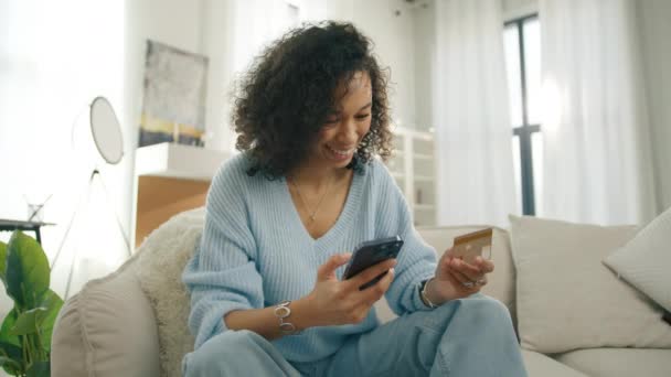 快乐的年轻的双族女人 深色皮肤 手握信用卡 在家里使用即时移动支付的多样化模型 在智能手机应用程序4K上购买的顾客微笑 — 图库视频影像