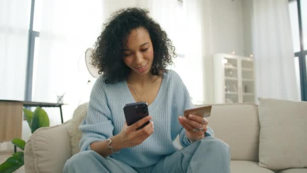 自宅でインスタントモバイル決済を使用した多様なモデル スマートフォンアプリ4Kで購入する笑顔のお客さん クレジットカードを保持するカーリーヘアとダークスキントーンを持つ幸せな若い偏見の女性 — ストック動画