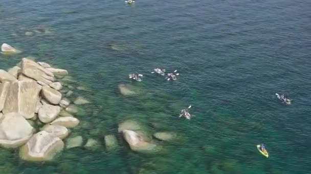 山岳湖で再生しているアクティブな観光客のグループを追跡するドローン 晴れた夏の晴れた湖岸 大きな丸い岩のタホ湖で透明な水に浮かぶクリアカヤックの空中撮影 — ストック動画