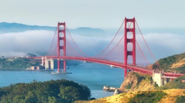 Arka planda beyaz bulut bulunan kırmızı Altın Köprü 'nün manzaralı görüntüsü. Sis Karl güneşli bir sabahta Pasifik Okyanusu 'ndan San Francisco Körfezi' ne sürükleniyor. Yeşil tepelerde yoğun sis var.