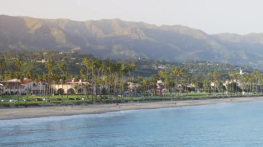 Santa Barbara tatil beldesi, Kaliforniya, ABD 'nin kıyı manzarası. Tropikal plajın palmiye ağaçlarıyla drone çekimi. Pasifik Okyanusu dalgaları kıyıda yuvarlanıyor. Dağları çevreleyen şehir silüeti 