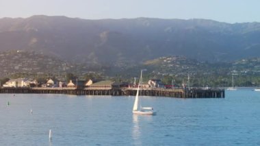 Stearns Wharf, Santa Barbara, California, ABD yakınlarında yüzen yelkenlilerin manzarası. Şehir limanındaki eski ahşap iskele. Gün batımında şehir binalarıyla dağlık tepelerin resimli görüntüleri, 4K görüntü. 