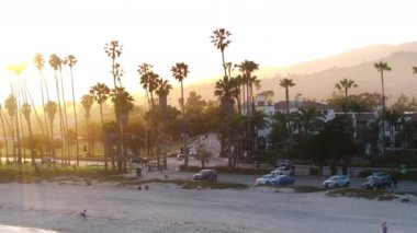 Santa Barbara şehrinin günbatımı manzarası, Kaliforniya, ABD. Trafikli büyük şehir hayatı. Kamerada parlayan güneş ışığı. Kumsalda eğlenen insanların robot resmi. Tatil konsepti, 4k görüntü 