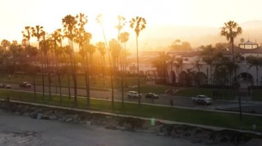 Santa Barbara silueti boyunca uçan insansız hava aracı ve günbatımında plaj, Kaliforniya, ABD. Pasifik kıyısındaki palmiye ağaçları boyunca güneş ışığı yanıp sönüyor. Şehirdeki konutların ve trafiğin hava görüntüsü 