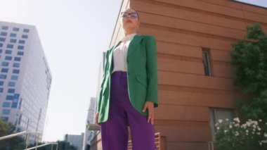 Düşük açılı, yavaş çekim. Moda düşkünü, zeki Amerikalı bir kadının modern şehir merkezinde hareketli bir arka planda gökdelenle kameraya bakarken kendine güveni tam bir şekilde bakması.