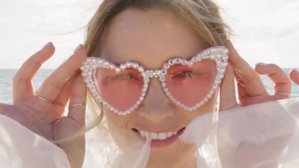 ピンクのメガネで若い女性を閉じ 海の背景のカメラにウインクします カメラを探しているグラマーなハートメガネの遊び心のある女性 ポートレートファッションガールがビーチでウィンクする ファッションモデルウィンクアイ — ストック動画