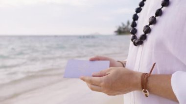 Yakından çekilen erkek eli, kabartmalı yemin kitabıyla birlikte. Geleneksel kukui fındık kolyeli Boho tarzı kıyafet içinde tanınmayan damat, sahil düğününde geline evlilik yemini kitabı okuyor.