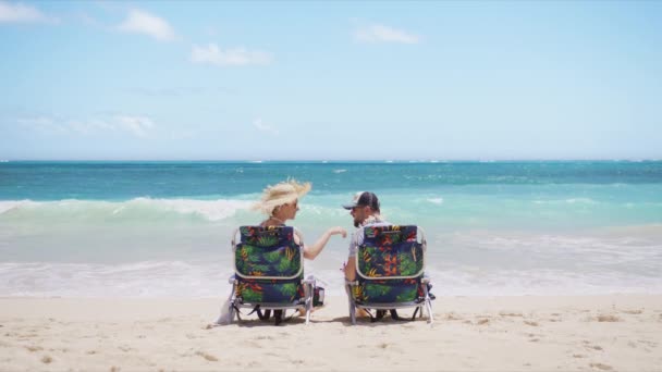 幸せな夫は夏の休日にトロピカルビーチに妻の花ハワイアンレイを置いています ビーチチェアの海に座っている男女 若いカップルは一緒にロマンチックなアウトドアライフスタイルの休暇を楽しむ — ストック動画